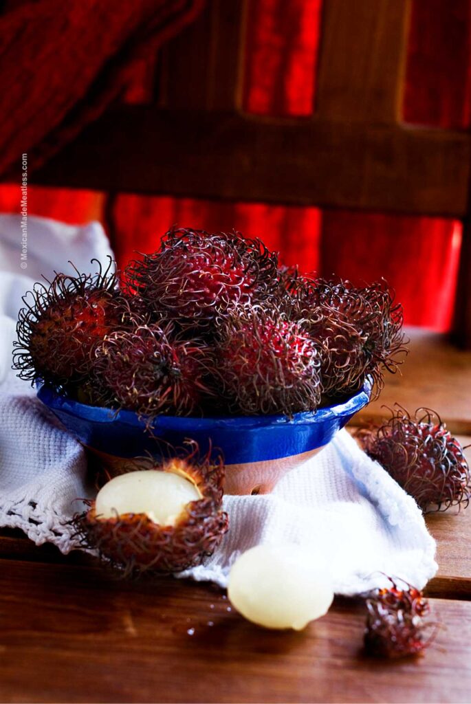A small blue bowl filled with rambutan fruit on a white cloth and on a wood table.