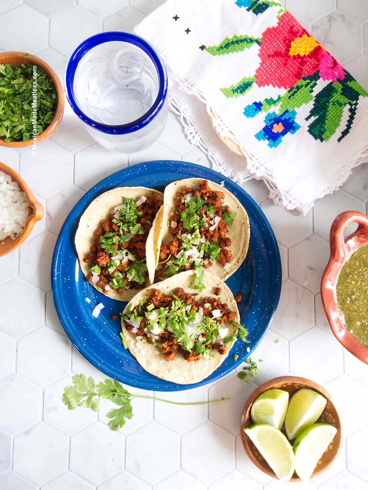 A blue plate with three vegan chorizo tacos topped with chopped onion and cilantro. On the side are quartered limes, salsa verde and a glass of water.
