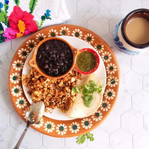 A view from above of a countertop with a decorative Mexican terracotta plate with a smaller white plate on top that's filled with vegan huevos con chorizo, corn tortillas, roasted salsa verde, and a small bowl filled with black beans. There's also a mug filled with cafe con leche.