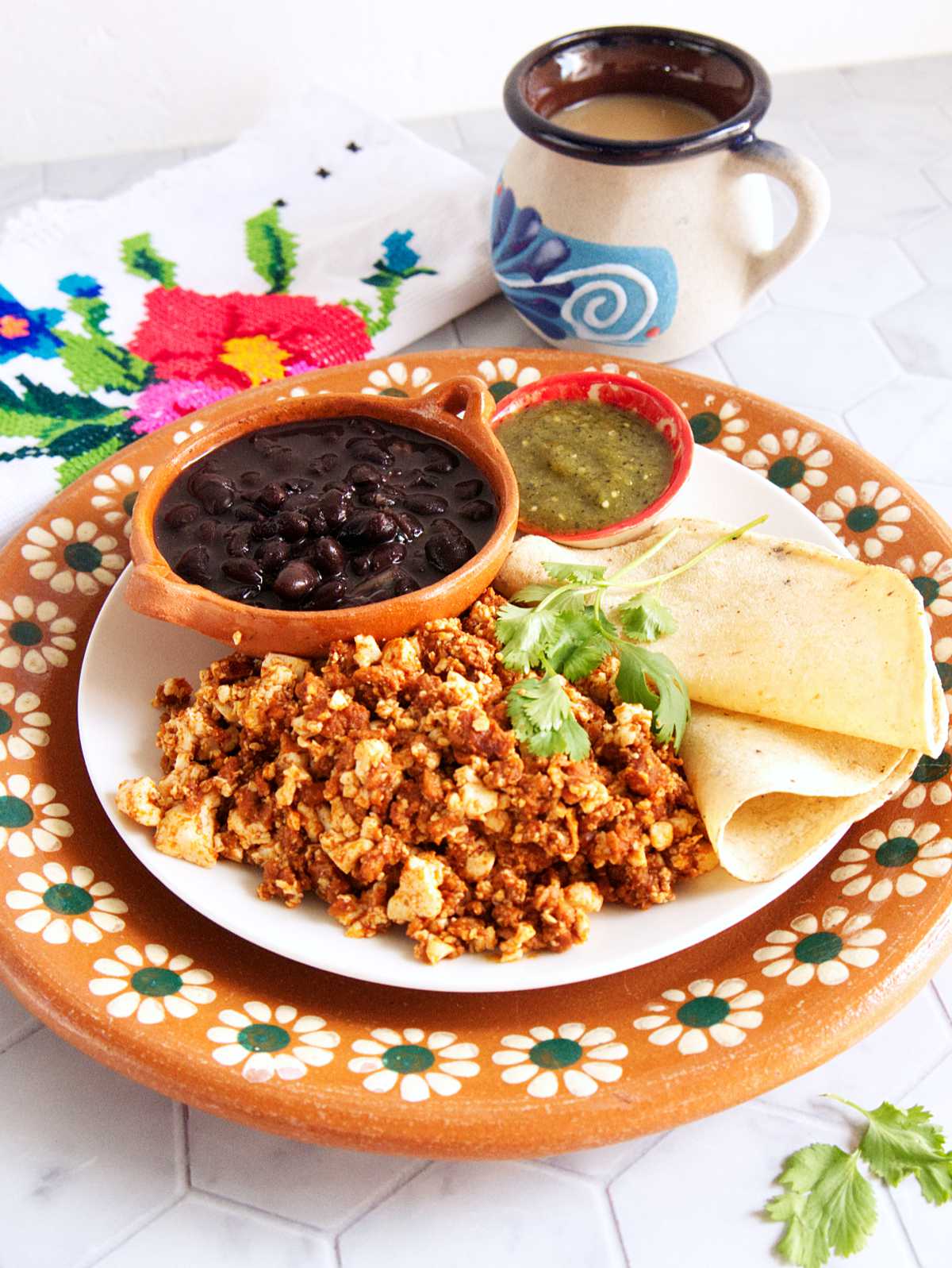 A decorative Mexican terracotta plate with a smaller white plate on top that's filled with vegan huevos con chorizo, corn tortillas, roasted salsa verde, and a small bowl filled with black beans. There's a mug in the background filled with cafe con leche.