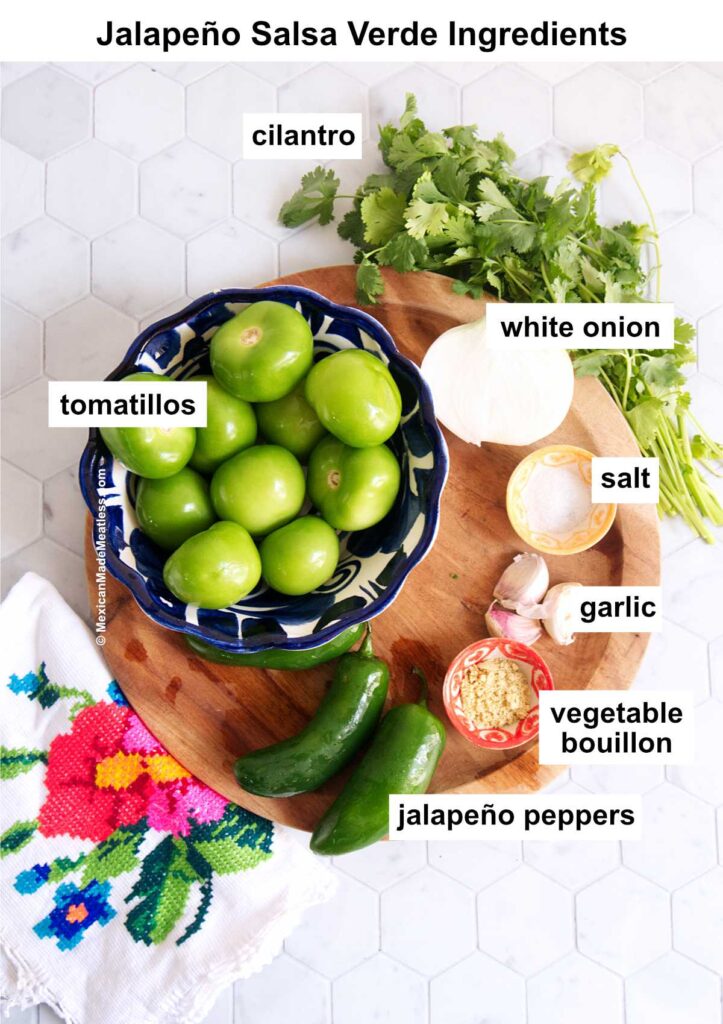 View from above of a kitchen countertop with a round wood platter that has some peeled tomatillos, raw jalapenos, onion, garlic, salt, vegetable bouillon powder and fresh cilantro.