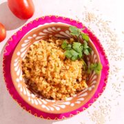 A vibrant dish of Mexican arroz made with brown rice and with peas garnished with cilantro in a decorative bowl, set on a pink and orange-patterned plate. Two tomatoes and scattered grains of rice are nearby on a light-colored surface.
