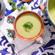 A bowl of Mexican crema de calabacitas soup garnished with fresh cilantro sits on a colorful patterned tablecloth. A spoon rests beside the bowl, and a pot of soup is partially visible, enhancing the traditional and vibrant design.