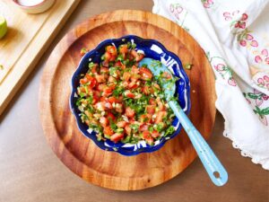 A colorful bowl of chunky salsa called pico de gallo made with chopped tomatoes, onions, cilantro, and jalapeños sits on a wooden board. A blue spoon rests inside the bowl. A decorative cloth with strawberry prints is to the side.