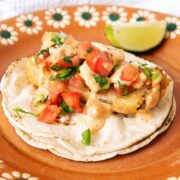 A dish with a vegan battered fish taco on a stack of corn tortillas, topped with diced tomatoes, cilantro, and creamy sauce. A lime wedge is on the side. The plate is decorated with a floral pattern.