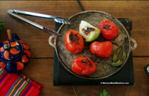 A metal comal on a hot plate holds roasted tomatoes, onion, and green chili for molcajete salsa. Metal tongs rest on the pan. Nearby are a colorful teapot and a striped cloth on a wooden table.