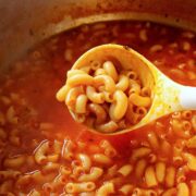 A white ladle scoops sopa de coditos—Mexican macaroni soup in a tomato-based broth—from a pot. The soup features elbow pasta and is seasoned with visible herbs.