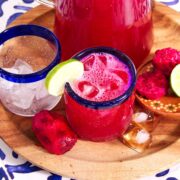 A wooden tray holds a glass of bright pink Agua de Tuna with ice and a lime wedge, an empty glass with ice, a pitcher of the same drink, halved prickly pears, lime halves, and ice cubes on a blue and white table.