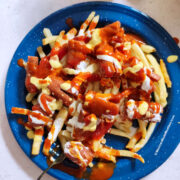 A blue plate filled with French fries topped with pieces of salchipulpos sausage, drizzled with ketchup, crema, hot sauce and mustard. A fork rests on the plate's edge.