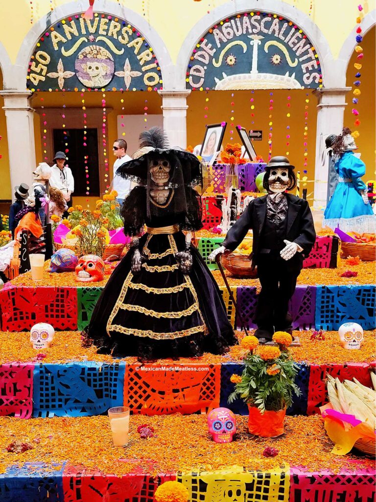 Day of the Dead altar adorned with papel picado, marigolds, candles, and skulls features Catrina and Catrin skeleton figures in elegant black attire beneath an archway reading “450 ANIVERSARIO DE AGUASCALIENTES.”.