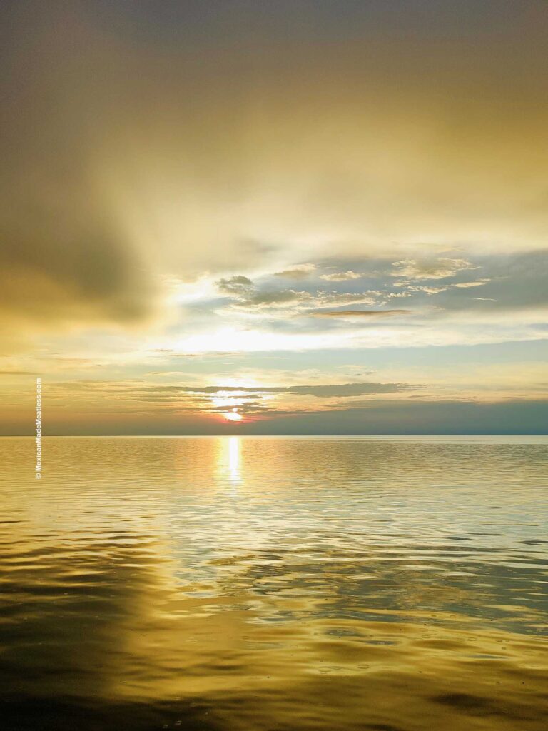 Golden sunset in Campeche, Mexico over a calm sea with soft clouds in the sky, sunlight reflected on the water, and a peaceful horizon.