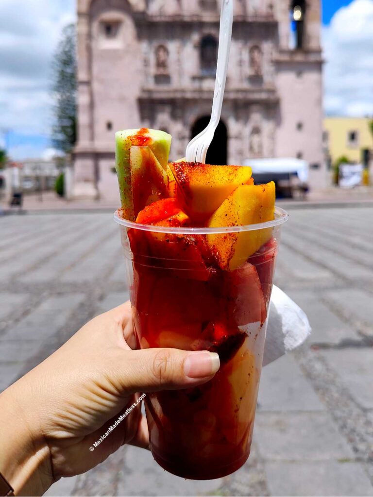 A woman's hand holds a clear cup filled with assorted fresh Mexican fruit slices topped with chili powder, with a plastic fork in the cup. A historic stone building and blue sky are visible in the blurred background.