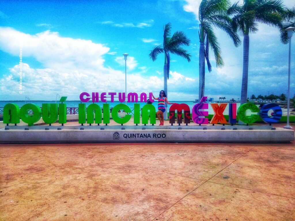 Colorful "Chetumal, aquí inicia México" sign with palm trees and a cloudy blue sky. Blogger Nancy Lopez poses behind the sign on the picturesque promenade in Quintana Roo, Mexico.