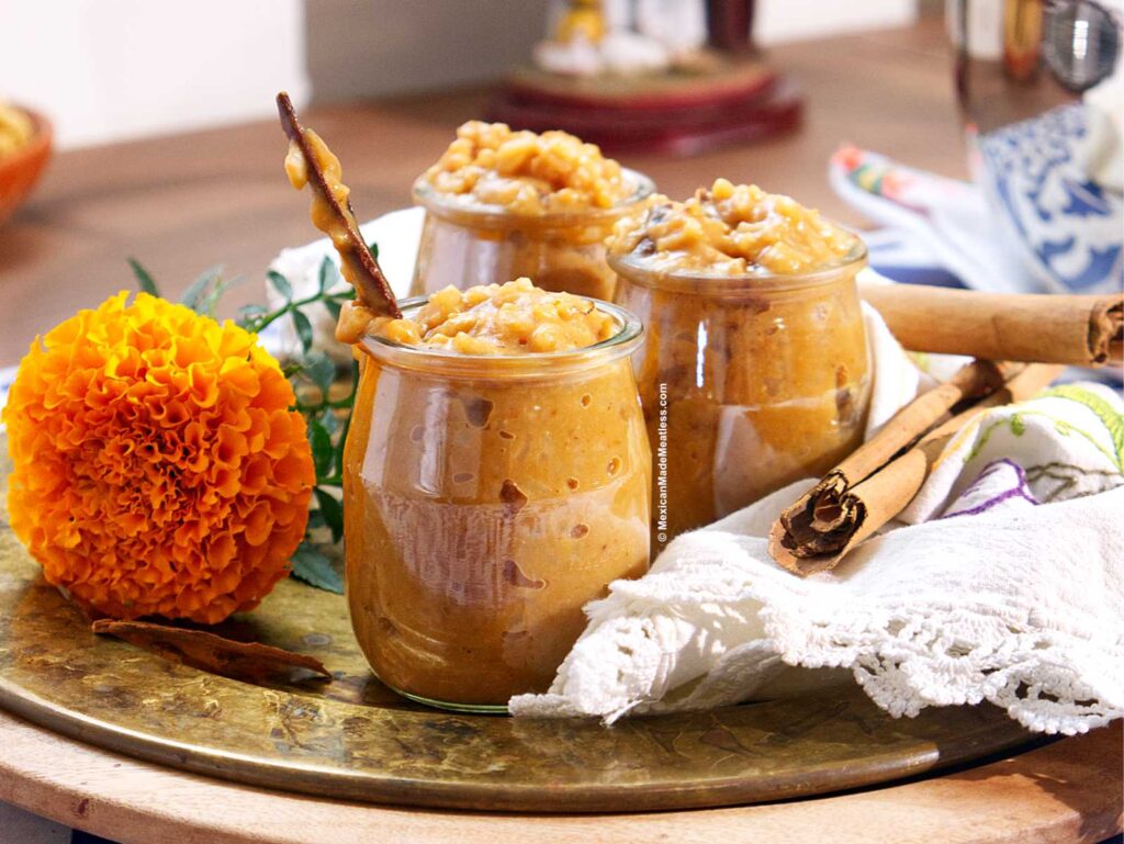 Three small glass jars filled with creamy, textured arroz con leche de calabaza are displayed on a round tray, accompanied by a vibrant orange marigold, cinnamon sticks, and a folded white napkin.