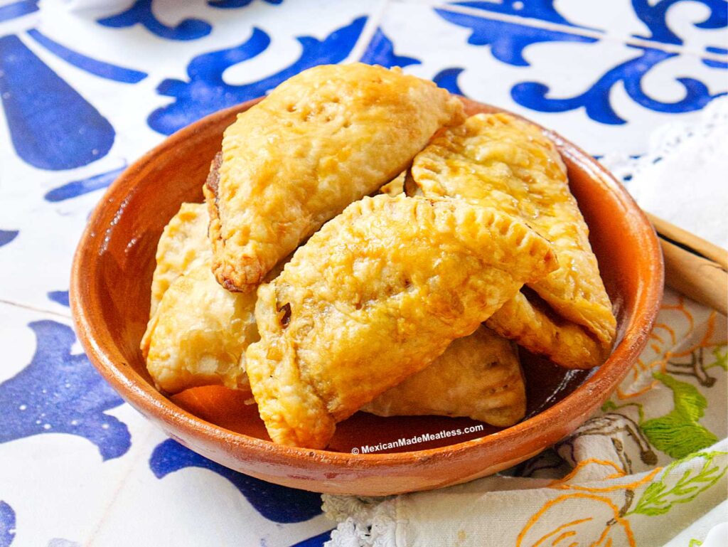 A clay bowl filled with golden-brown, flaky Mexican pumpkin empanadas sits on a colorful tiled surface next to a patterned cloth napkin.