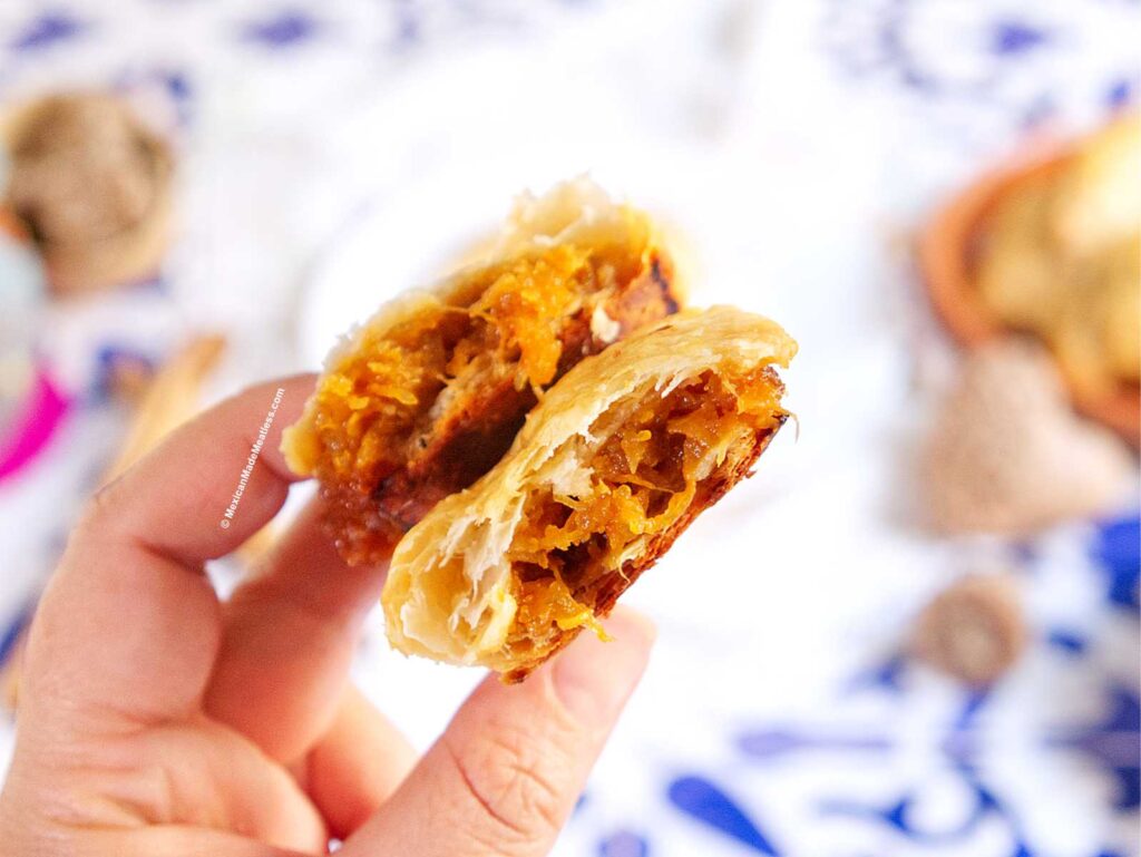 A hand holds an empanada split in half, revealing a golden, flaky crust and a spiced, pumpkin filling. The background is blurred with hints of blue and white patterns.