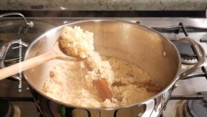 A saucepan on a stove contains cooked rice with a cinnamon stick, and a wooden spoon scoops some rice from the pot. Steam rises from the rice, indicating it is hot and freshly cooked.
