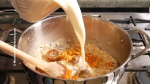 Milk is being poured from a measuring cup into a pot of rice, pumpkin puree, piloncillo, and other ingredients, while a wooden spoon stirs the mixture on a stove.