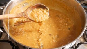 A pot of creamy pumpkin arroz con leche with piloncillo simmers on the stove, while a wooden spoon lifts a portion to reveal its thick, textured consistency and golden-brown hue.