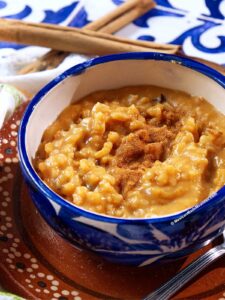 A blue and white bowl filled with creamy pumpkin arroz con leche, sweetened with piloncillo and topped with ground cinnamon, sits on a brown plate with a spoon. Cinnamon sticks and a blue-patterned cloth are visible in the background.