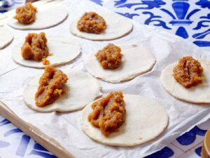 Round pastry dough circles on parchment paper, each topped with a spoonful of empanada filling, arranged on a wooden board against a blue and white patterned background.