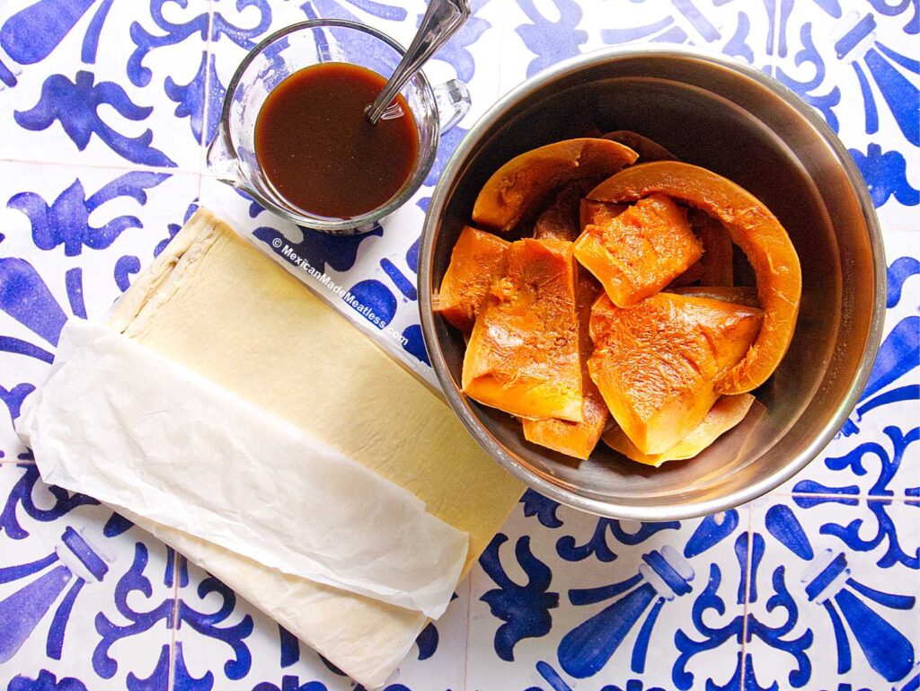A metal bowl with cooked pumpkin pieces, a glass cup of piloncillo syrup, and a folded sheet of pastry on white and blue patterned tiles.