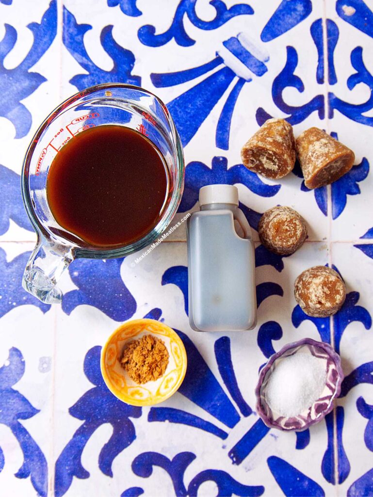 A top-down view of ingredients on a blue and white patterned tile: a measuring cup with piloncillo syrup, a small bottle of vanilla extract, piloncillo cones, a small bowl of cinnamon, and a small bowl of white salt.