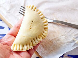 A hand holds an uncooked pumpkin empanada with crimped edges and small fork holes on top. A fork and parchment paper are visible in the background.