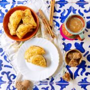 Three pumpkin empanadas made with puff pastry rest on a white plate, with more in a bowl. Cinnamon sticks, a colorful mug of coffee, piloncillo sugar pieces, and a clay jug sit atop decorative blue-and-white tiles.