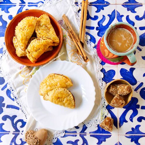 Three pumpkin empanadas made with puff pastry rest on a white plate, with more in a bowl. Cinnamon sticks, a colorful mug of coffee, piloncillo sugar pieces, and a clay jug sit atop decorative blue-and-white tiles.