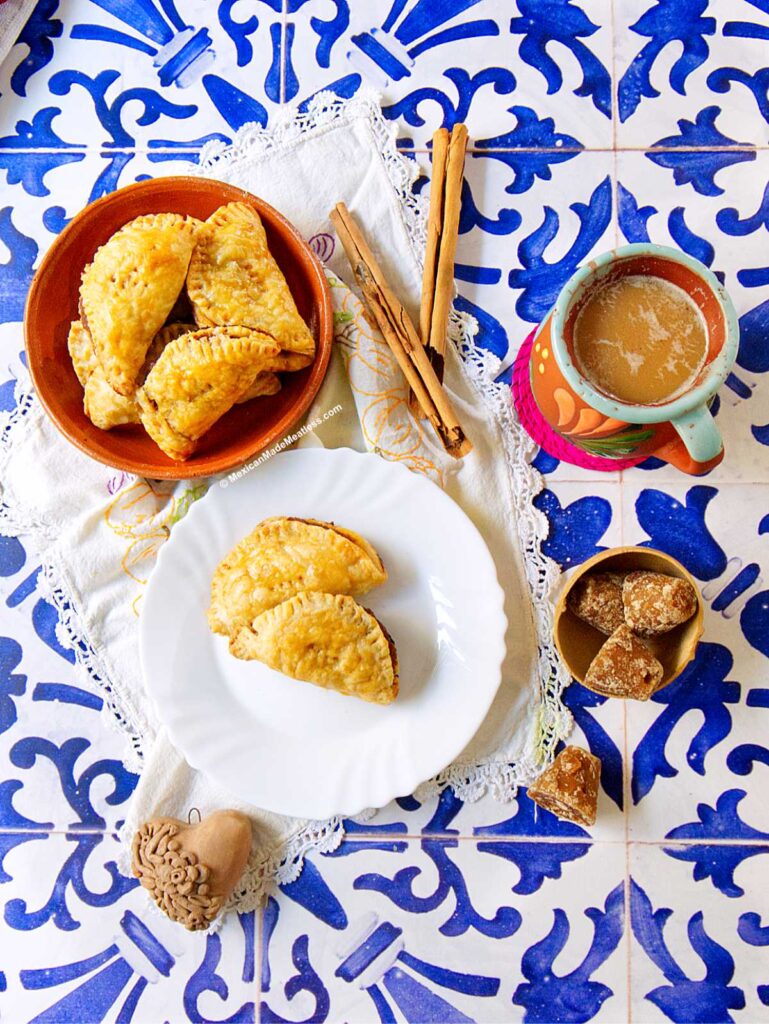 Three pumpkin empanadas made with puff pastry rest on a white plate, with more in a bowl. Cinnamon sticks, a colorful mug of coffee, piloncillo sugar pieces, and a clay jug sit atop decorative blue-and-white tiles.