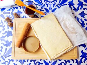 A sheet of puff pastry on parchment paper sits on a wooden board with a rolling pin, round tin lid, small jar of piloncillo syrup, and chunks of piloncillo on a blue and white patterned surface.