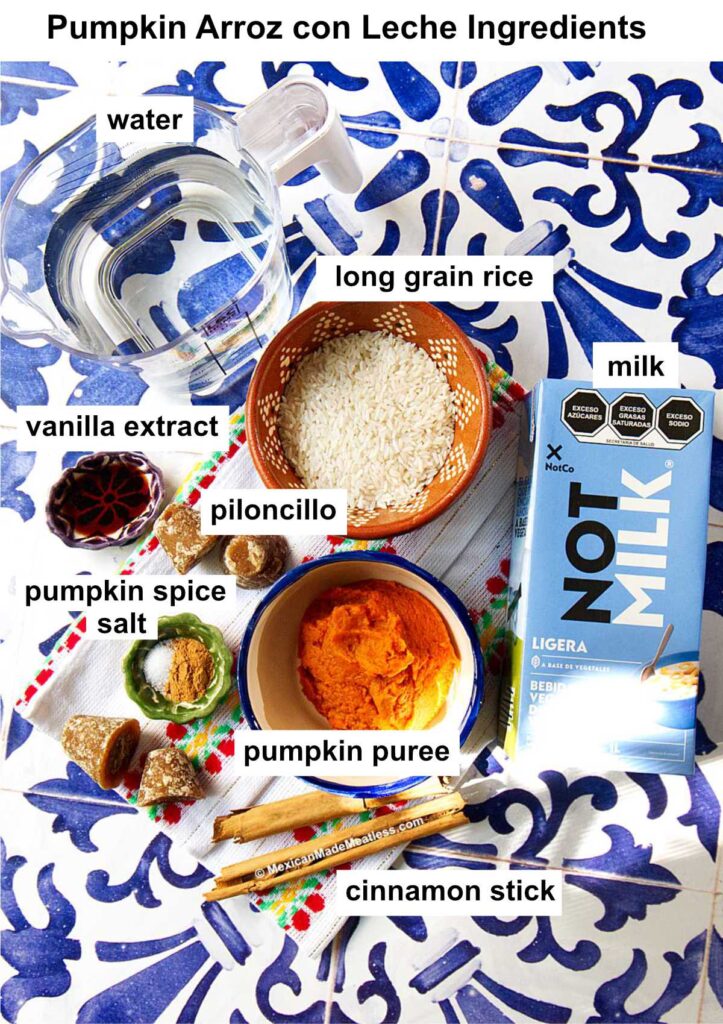 Overhead view of pumpkin arroz con leche ingredients: water, long grain rice, milk, vanilla extract, piloncillo, pumpkin spice, salt, pumpkin puree, and a cinnamon stick arranged on a blue and white tile.