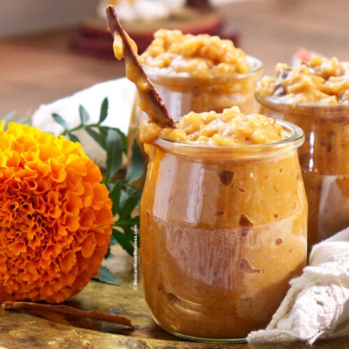 Three glass jars filled with creamy vegan pumpkin arroz con leche, garnished with a cinnamon stick, are placed on a tray next to a vibrant orange marigold flower and a white cloth.