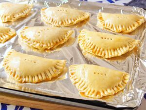 Eight uncooked empanadas arranged on a foil-lined baking sheet. The pastries are golden and neatly crimped along the edges, ready to be baked.