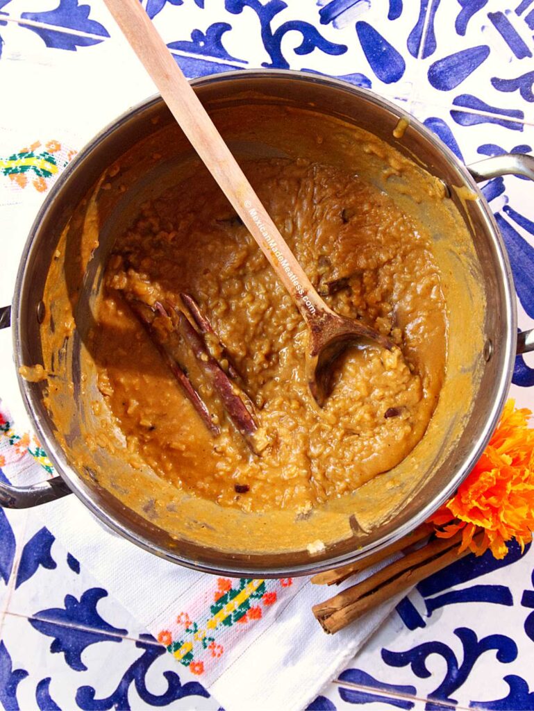 A pot of creamy Mexican pumpkin rice pudding with a wooden spoon sits atop a colorful patterned tablecloth, accented by whole cinnamon sticks, an orange marigold flower, and extra cinnamon beside the pot.