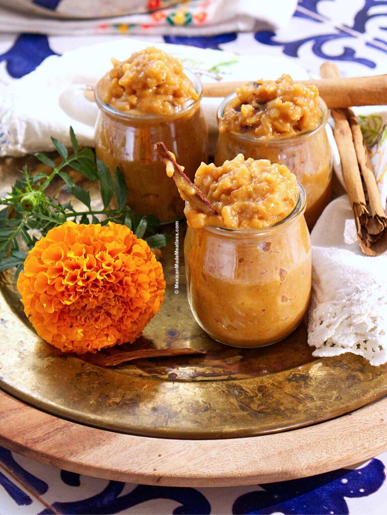 Three small glass jars filled with creamy, dairy-free pumpkin arroz con leche are placed on a brass tray next to cinnamon sticks, a bright orange marigold flower, and a white napkin with colorful embroidery.