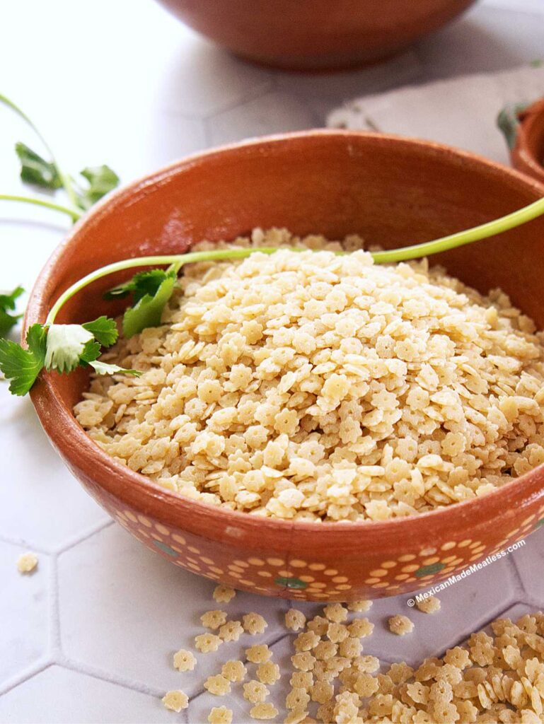 A brown, flower-patterned bowl filled with uncooked star pasta sits on a white surface, garnished with a sprig of fresh cilantro. Some star-shaped pasta pieces are scattered nearby.
