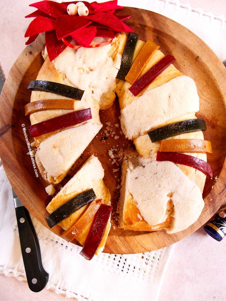 A sliced Rosca de Reyes, the traditional round sweet bread decorated with colorful candied fruit, sits on a wooden platter. A festive red felt decoration tops the Rosca de Reyes, with a knife resting beside the platter.