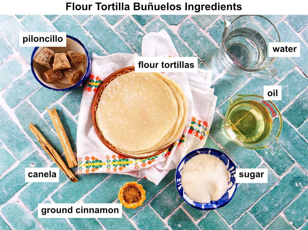 A flat lay of labeled ingredients for flour tortilla buñuelos: flour tortillas on a towel, bowls of piloncillo, sugar, ground cinnamon, whole cinnamon sticks (canela), and glasses of water and oil on a green tile surface.