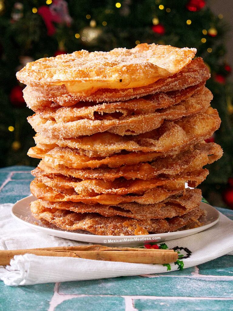 A tall stack of easy Mexican buñuelos, golden and crispy, dusted with cinnamon sugar sits on a plate, with a festive, blurred Christmas tree in the background and cinnamon sticks on the table beside the plate.