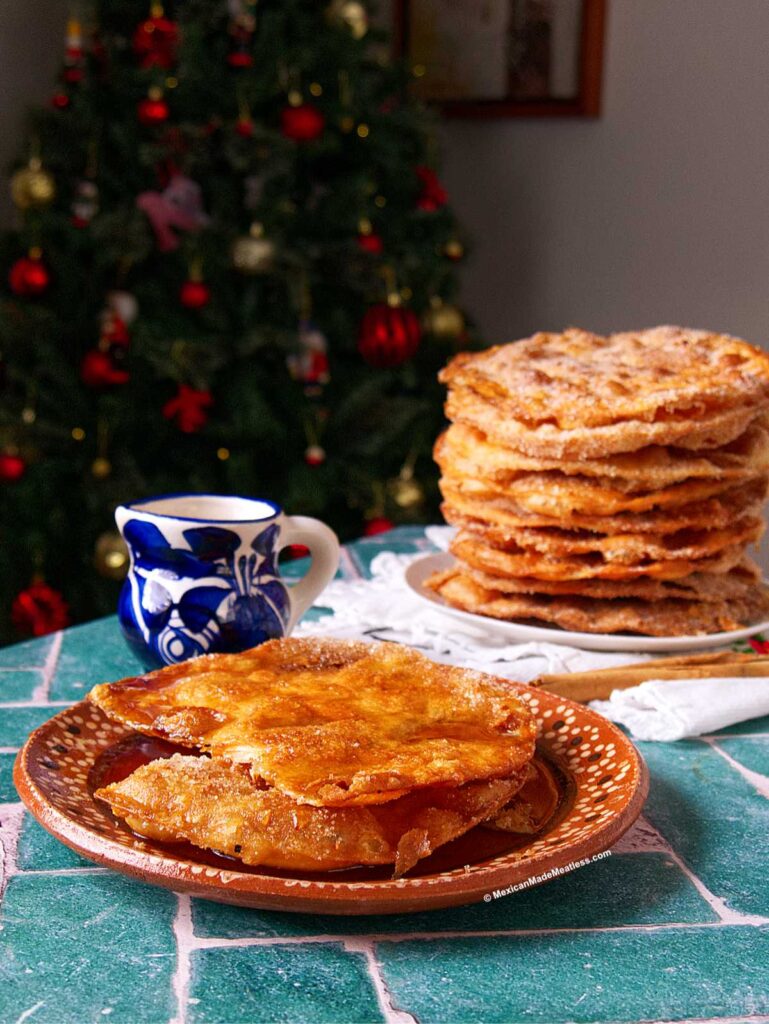 A plate with two golden, crispy buñuelos with piloncillo syrup sits on a green table, next to a blue and white cup. In the background, there is a stack of more buñuelos and a decorated Christmas tree.