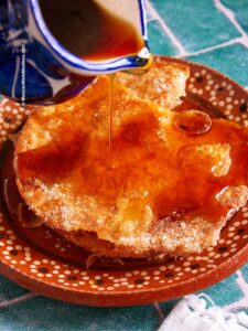 A close-up of a Mexican buñuelos fritter with piloncillo syrup being poured over it, resting on a decorative clay plate. The golden brown treat is sprinkled with sugar, set against the backdrop of green tile.