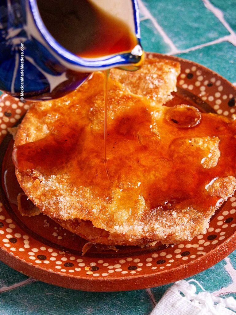 A close-up of a Mexican buñuelos fritter with piloncillo syrup being poured over it, resting on a decorative clay plate. The golden brown treat is sprinkled with sugar, set against the backdrop of green tile.