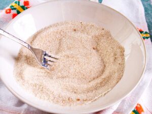 A white bowl filled with cinnamon sugar being mixed with a fork, placed on a patterned white cloth.