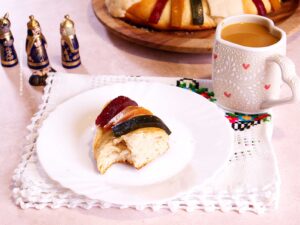 A slice of Rosca de Reyes topped with candied fruit sits on a white plate beside a mug of coffee. The festive bread rests on an embroidered cloth, with figurines and more Rosca de Reyes in the background.