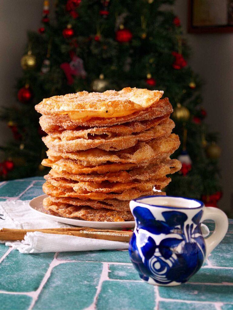 A tall stack of sugar-coated buñuelos, made from an easy buñuelos recipe, sits on a plate beside a blue and white ceramic mug filled with miel de piloncillo, with a decorated Christmas tree in the blurred background.