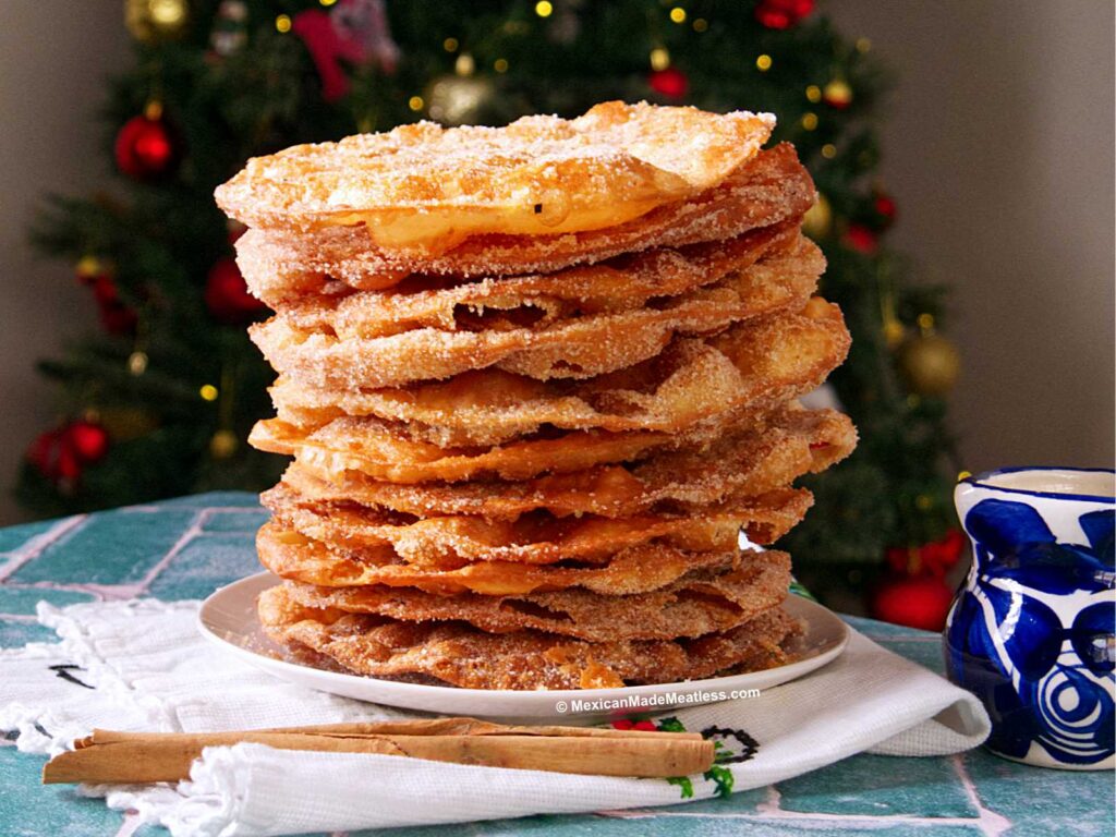 A tall stack of round, crispy buñuelos sprinkled with sugar sits on a white plate. In the background, a decorated Christmas tree is visible. Cinnamon sticks and a blue-patterned mug are nearby on the table.