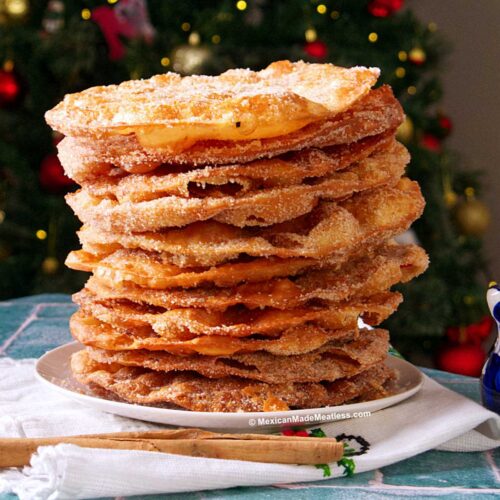 A tall stack of round, crispy buñuelos sprinkled with sugar sits on a white plate. In the background, a decorated Christmas tree is visible. Cinnamon sticks and a blue-patterned mug are nearby on the table.