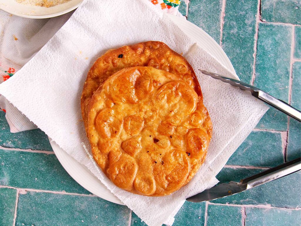 Two pieces of golden-brown fried bunuelos rest on paper towels on a white plate, with metal tongs nearby, atop a green-tiled surface.